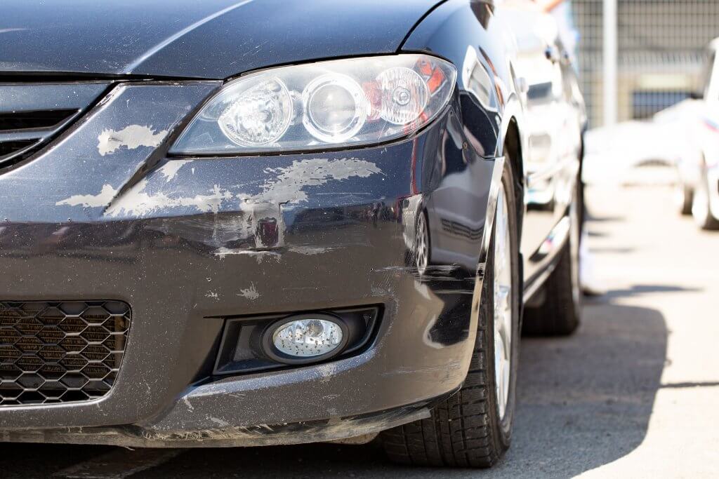 Nahaufnahme der vorderen Stoßstange eines schwarzen Autos mit Kratzern und Lackschäden, geparkt am Straßenrand