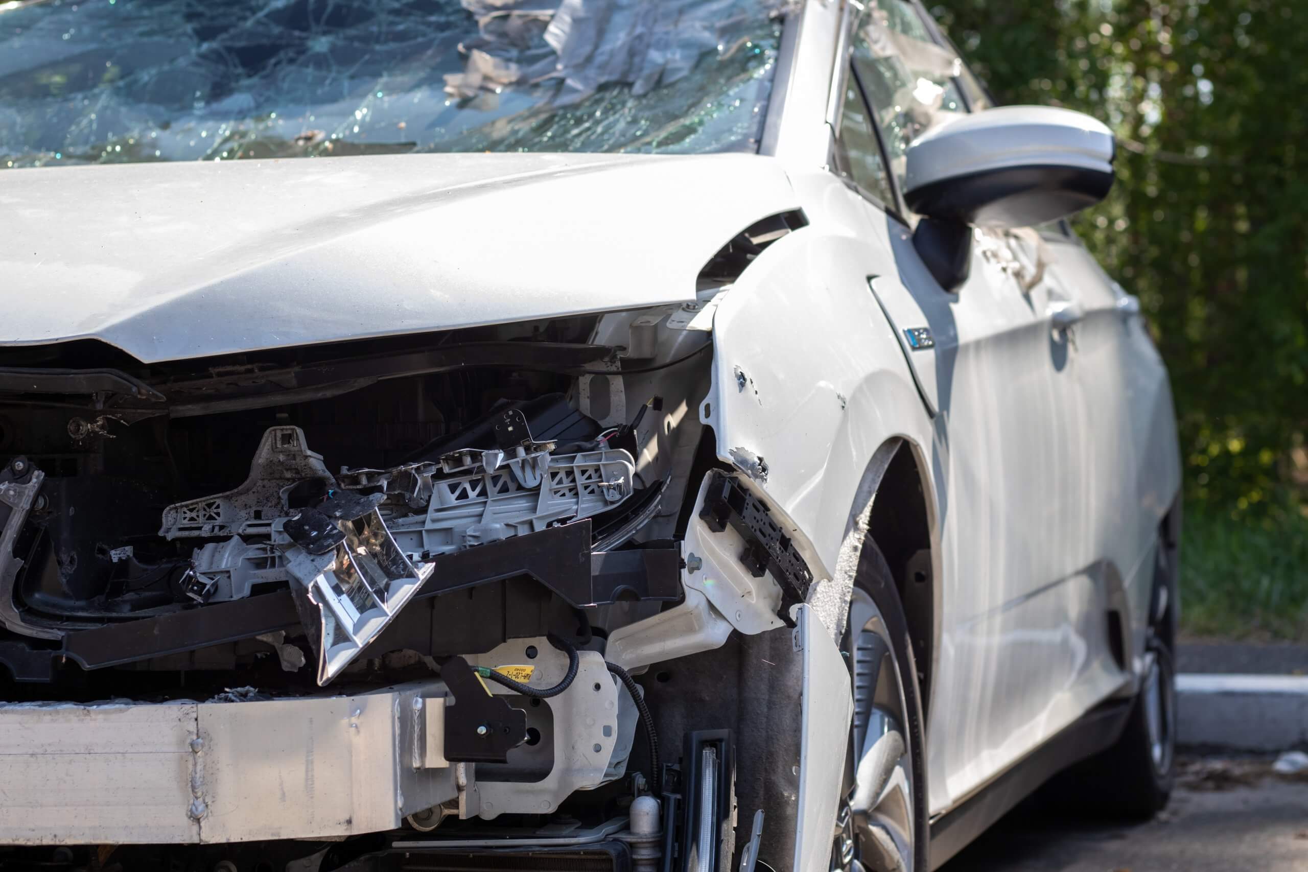Ein silbernes Auto mit zerstörter Front und zerbrochener Windschutzscheibe steht neben Grünfläche