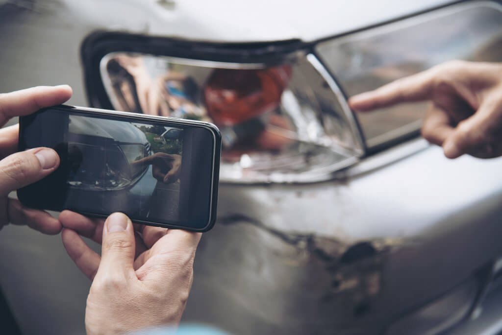 Eine Person fotografiert einen beschädigten Autoscheinwerfer, während eine andere Hand auf Unterschiede für die Reparaturkostenkalkulation zeigt