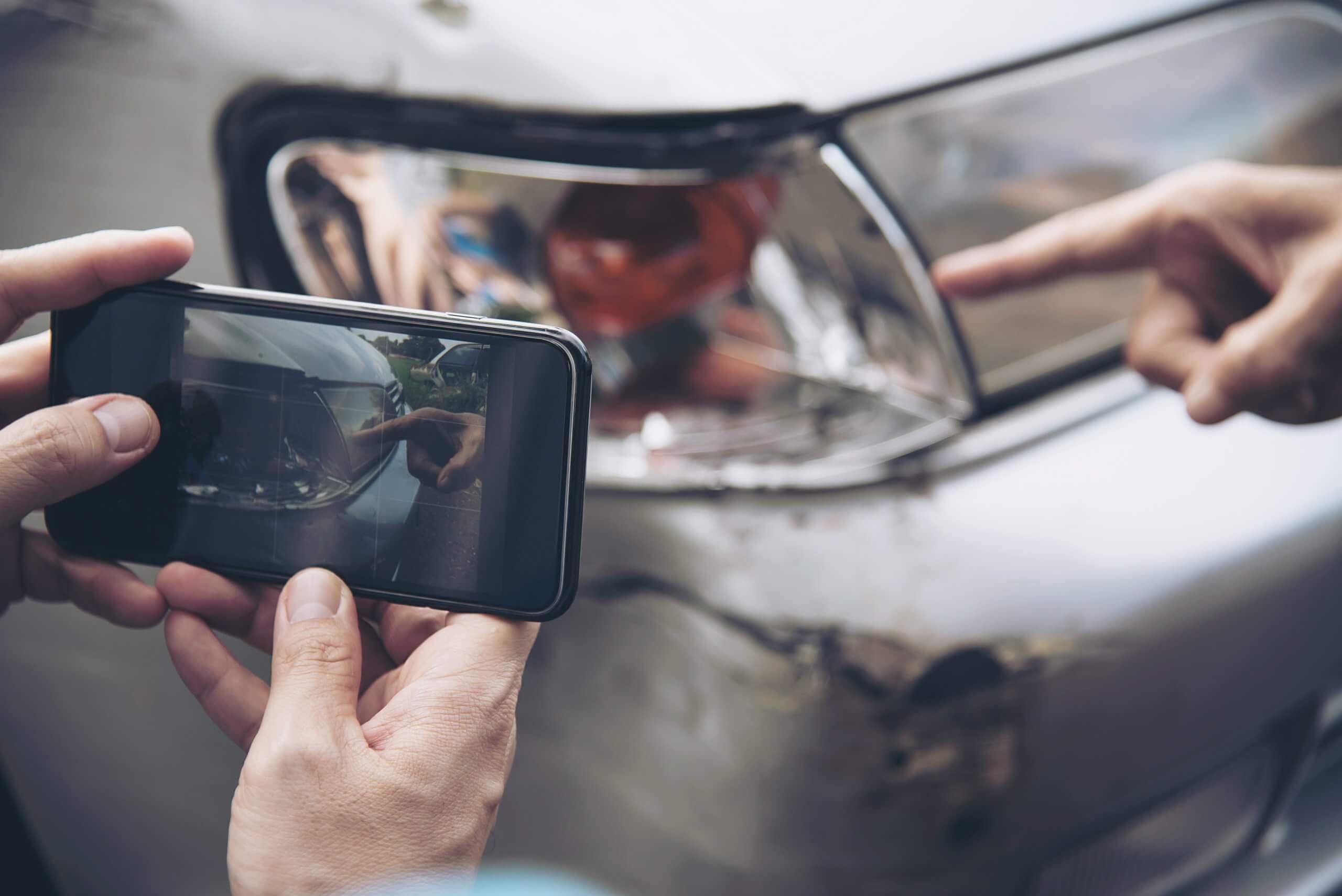 Eine Person fotografiert einen beschädigten Autoscheinwerfer, während eine andere Hand auf Unterschiede für die Reparaturkostenkalkulation zeigt